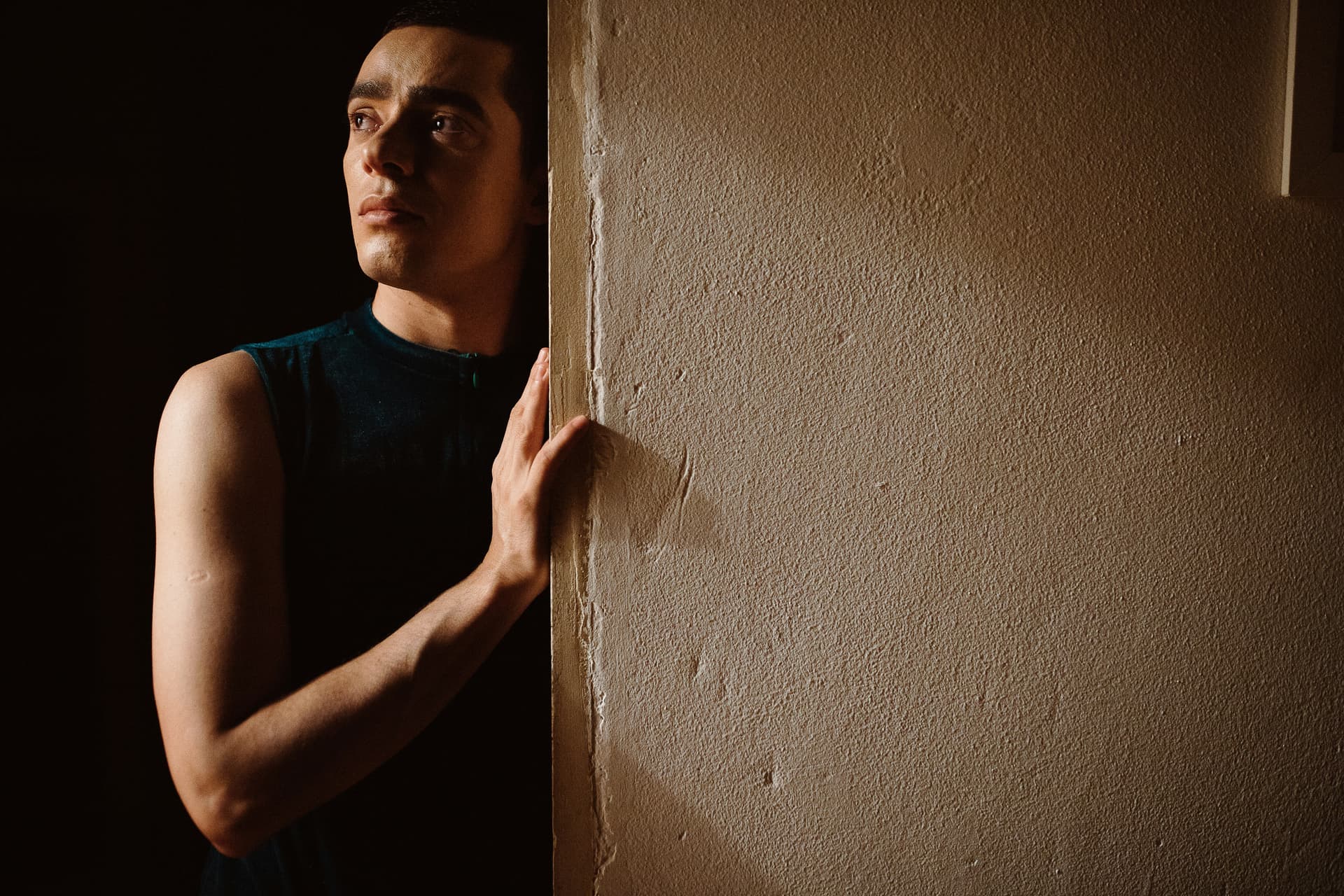 Person in a sleeveless top leaning against a textured wall in a dimly lit room.