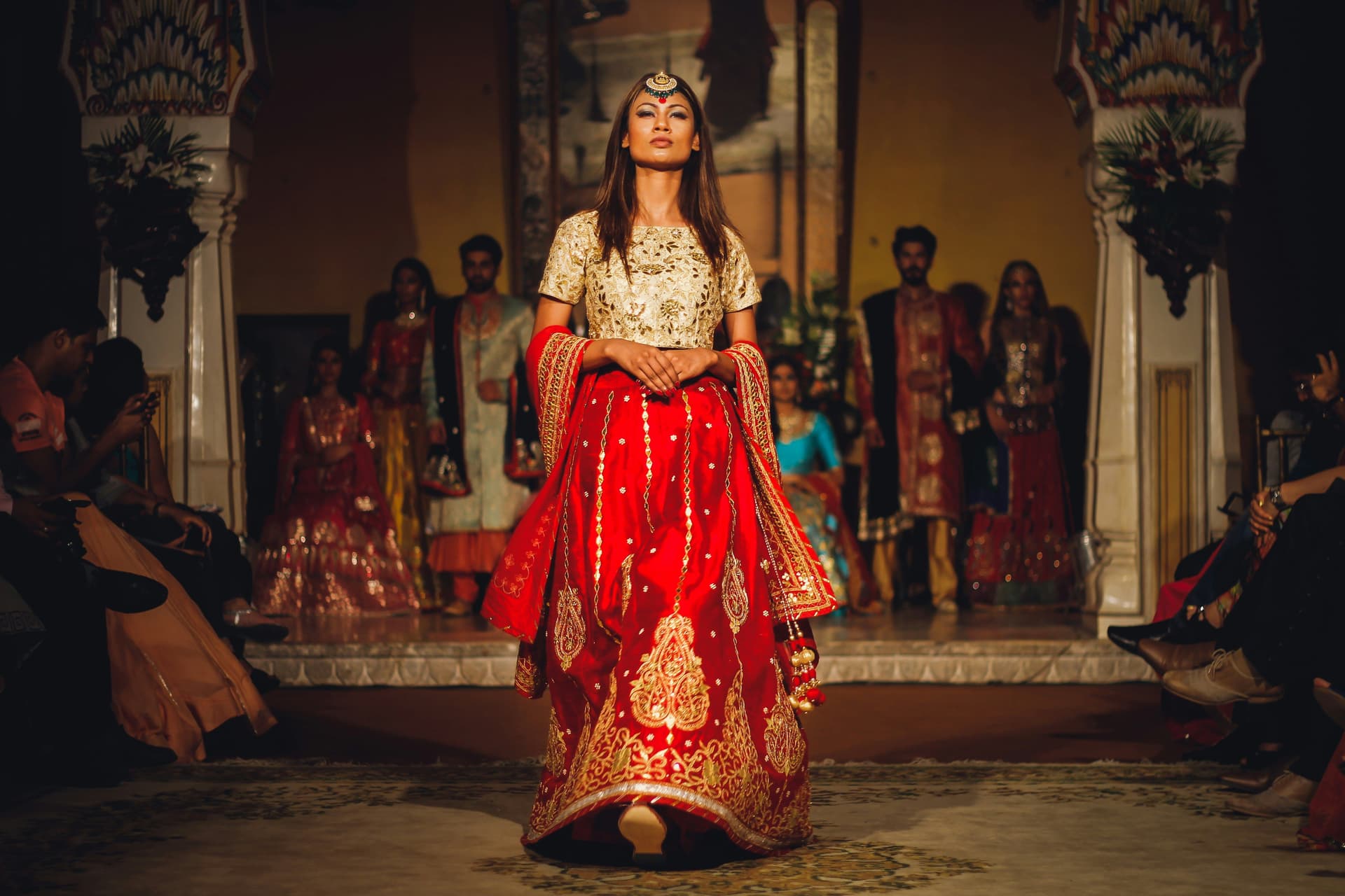 Person in ornate red and gold traditional attire on a stage with audience on sides.